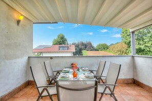 Un balcón techado con mesa lista para comer, sillas y vistas a una casa y árboles.