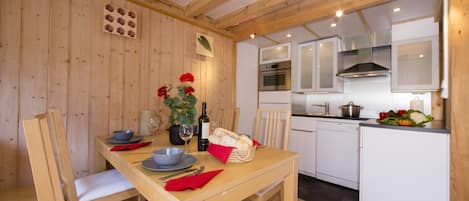 Une table à manger en bois avec des chaises, un coin cuisine avec des meubles blancs et un plafond en bois.