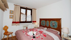 A bedroom with a checkered bedspread, a wooden headboard with floral artwork, a wooden chair, a radiator, and a window with curtains. - Vista Livigno, Livigno, Italy (Livigno)