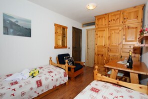 Una habitación con dos camas individuales, un armario de madera, una silla, una mesa con una botella y un vaso, y un cuadro en la pared.