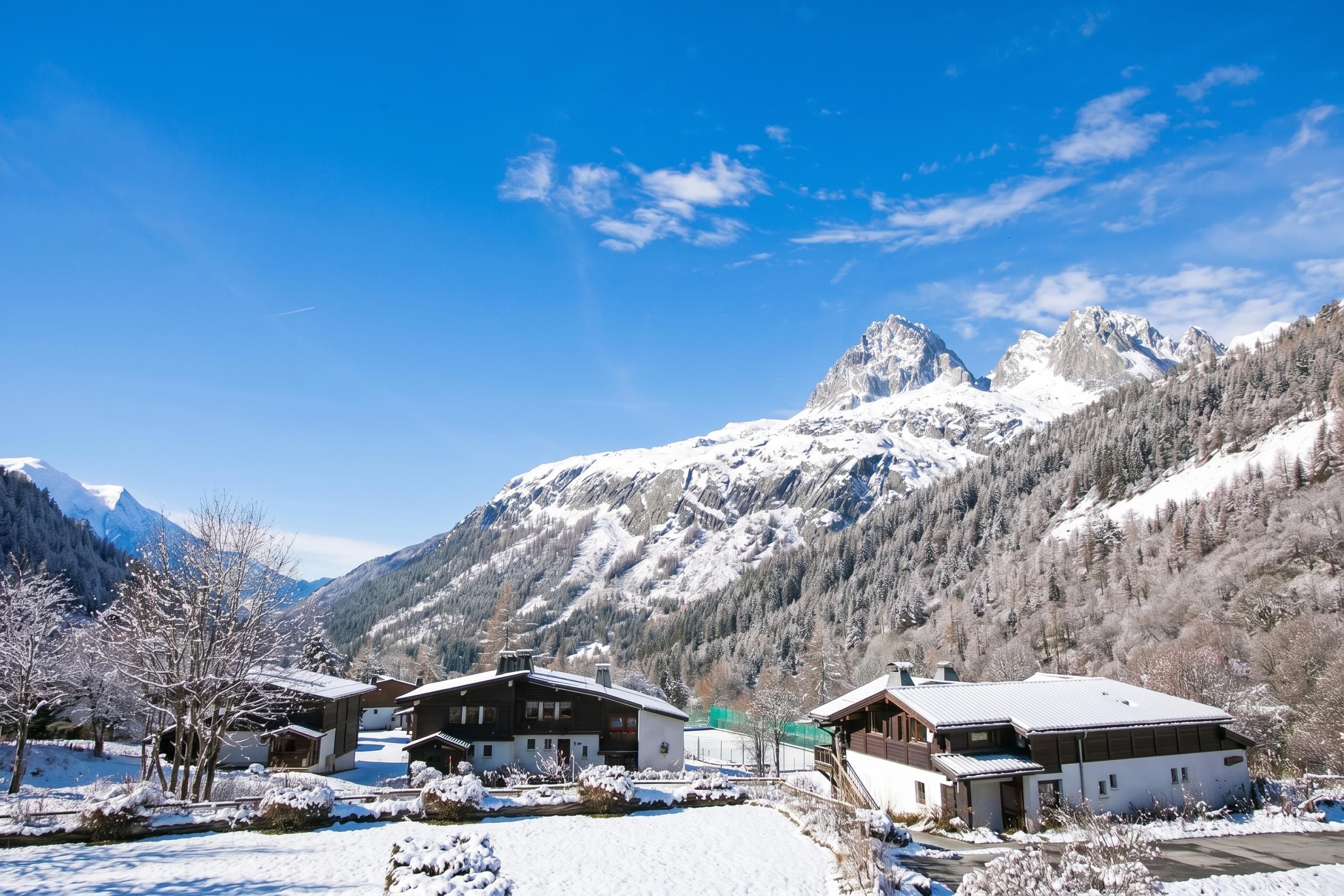 A mountain resort with chalet-style buildings, a clear sky, and snow-covered peaks in the background.