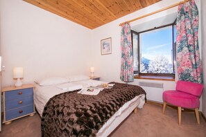 Quarto com cama, mesas de cabeceira, uma cadeira, janela com cortinas e um quadro na parede.