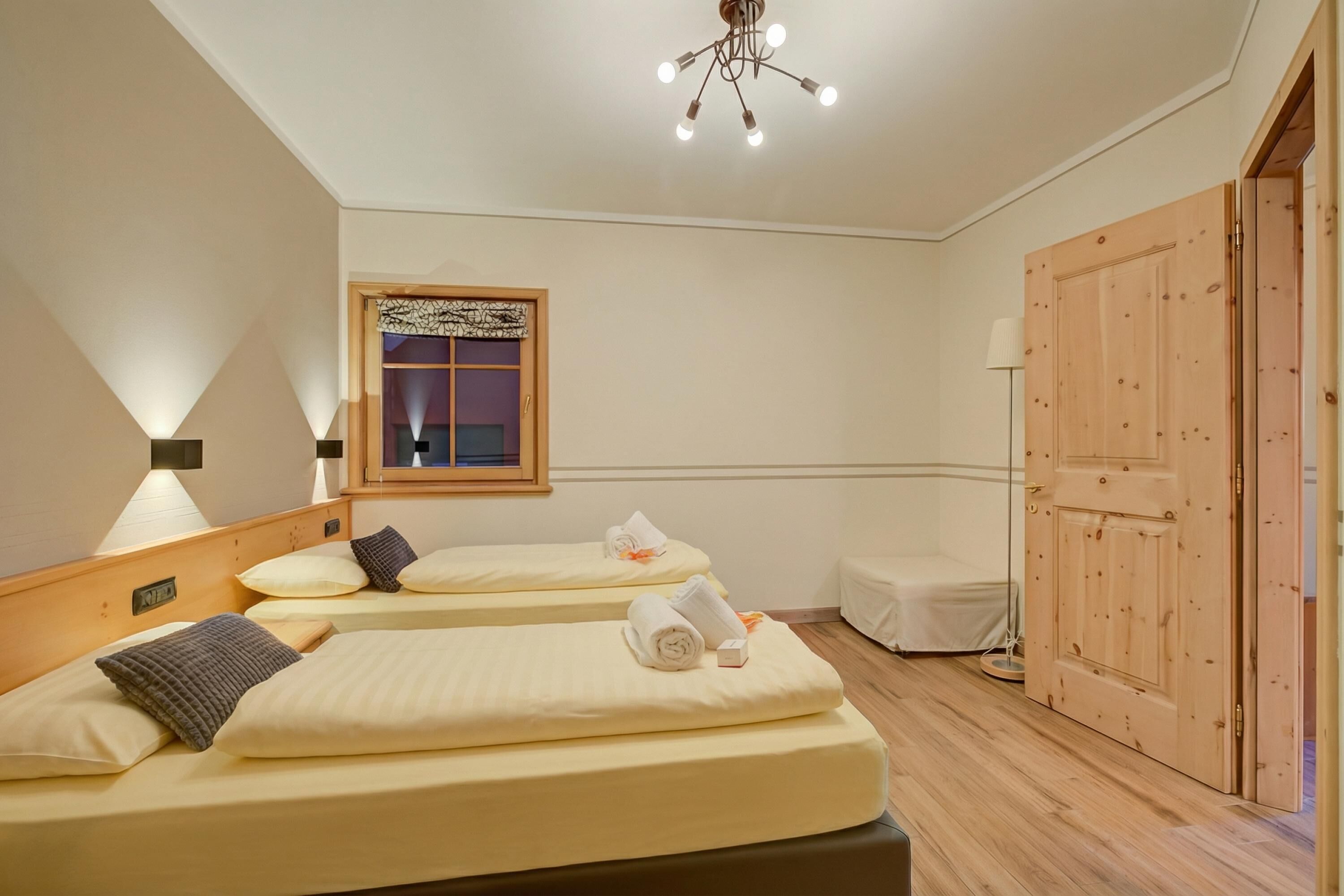 A hotel room with two beds, a wooden door, a window with a curtain, a ceiling light fixture, and a bedside table with a lamp.