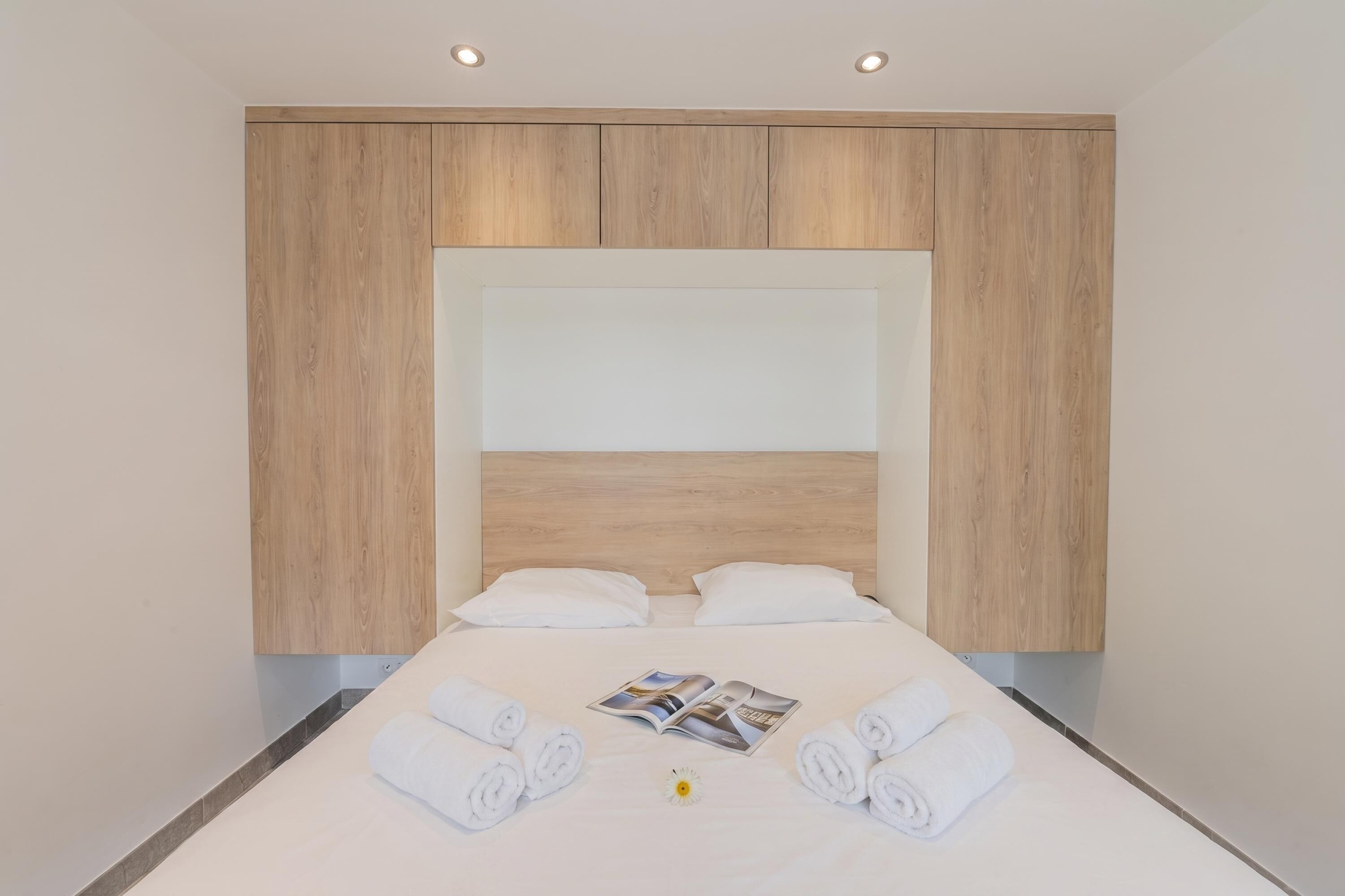 A hotel room with a bed, two rolled towels, a magazine, and a small flower.