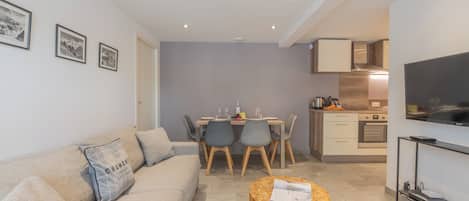 Una sala de estar moderna con un sofá, una mesa de centro redonda, un área de comedor con sillas, una cocina con armarios y un televisor de pantalla plana montado en la pared.