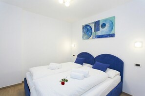 A neatly made bed with white linens and blue headboard, a single red rose, and abstract wall art.
