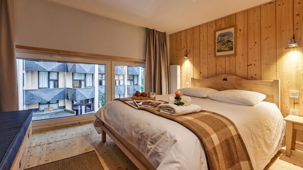 A bedroom with a large window, wooden walls, a bed with a blanket, and a nightstand with a lamp. - Grand Roc C122 280m from Slopes & Parking, Argentière (Chamonix), France (Argentière (Chamonix))