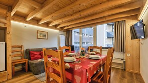 A dining area with a table set for four, wooden chairs, a red tablecloth, and a television mounted on the wall. - Grand Roc C122 280m from Slopes & Parking, Argentière (Chamonix), France (Argentière (Chamonix))