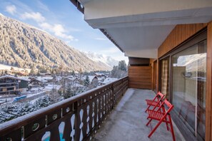 A balcony with a wooden railing, two red chairs, and a view of snow-covered mountains and buildings.