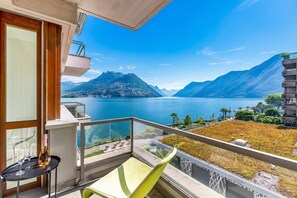 A balcony with a view of a lake and mountains, a table with glasses and a bottle, and a green chair. - Paradiso Incanto Lake View Apartment, Paradiso (Lugano), Switzerland (Paradiso (Lugano))