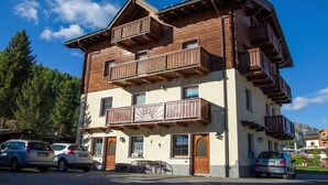 A multi-story wooden building with balconies, parked cars, and a mountain backdrop. - Italia 1 Ski in- Ski out 50m, Livigno, Italy (Livigno)