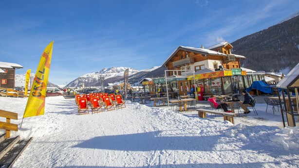 Een besneeuwde buitenruimte met houten banken en tafels, een restaurant en een berg op de achtergrond.