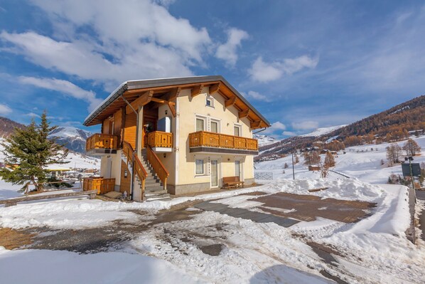 A chalet-style building with a wooden balcony, a staircase leading to the entrance, and a snowy landscape in the background.