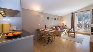 A modern kitchen with a fruit bowl, dining table, and chairs. There is a living area with a sofa and a window. - Cham Sud Forclaz504 - 1BR w/ Balcony sleeps 4, Chamonix, France (Chamonix)