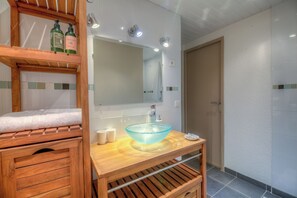 Uma casa de banho com toucador de madeira, lavatório em vidro, espelho e prateleira com artigos de higiene pessoal.
