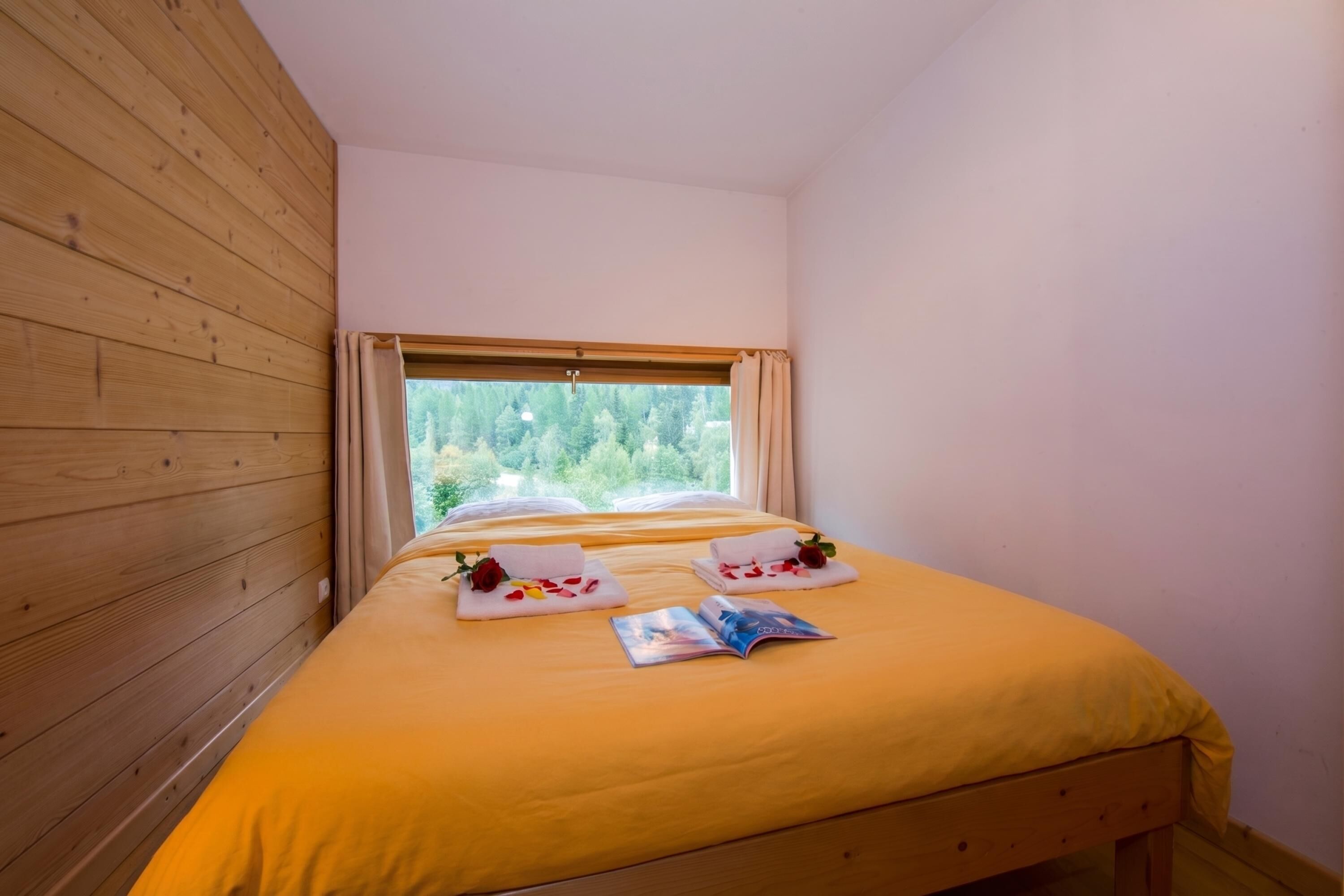 Una habitación con pared de madera, una cama con colcha amarilla, una ventana con cortinas y vistas a la vegetación exterior.