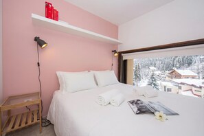A bedroom with a bed, pillows, a bedside table, a shelf with books, and a window with a snowy view. - Grand Roc C303 280m from Slopes, View, Parking & WiFi, Argentière (Chamonix), France (Argentière (Chamonix))