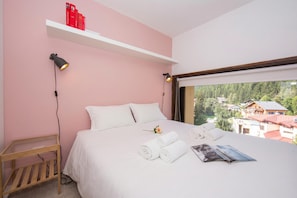 Un dormitorio con cama, almohadas, una mesita de noche, lámpara, ventana con vistas y una estantería con libros.