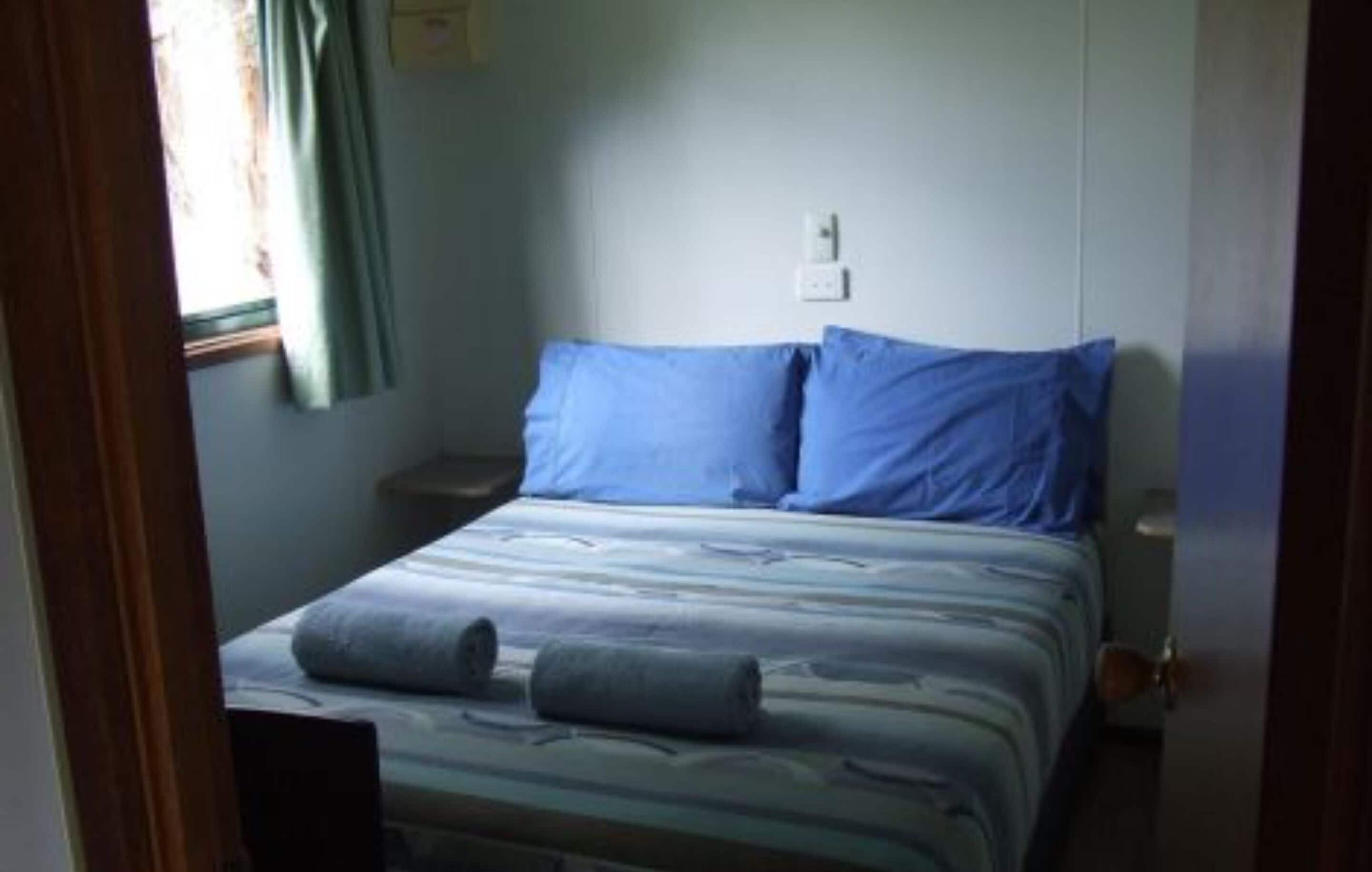 Un dormitorio con cama, dos almohadas, una mesita de noche, una ventana con cortinas y un enchufe.