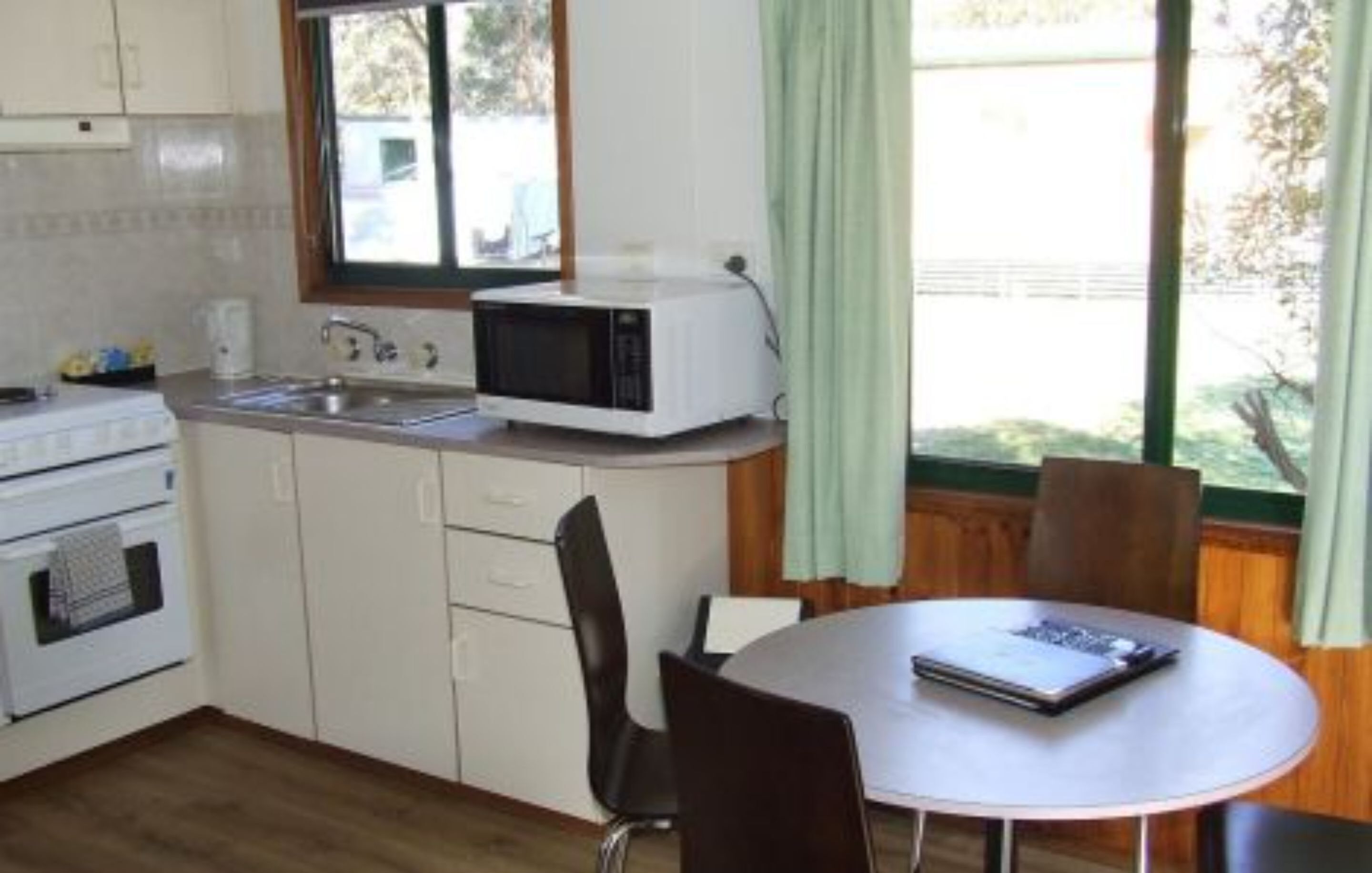 A compact kitchen with a microwave, sink, and stove, a dining table with chairs, and a window with curtains.