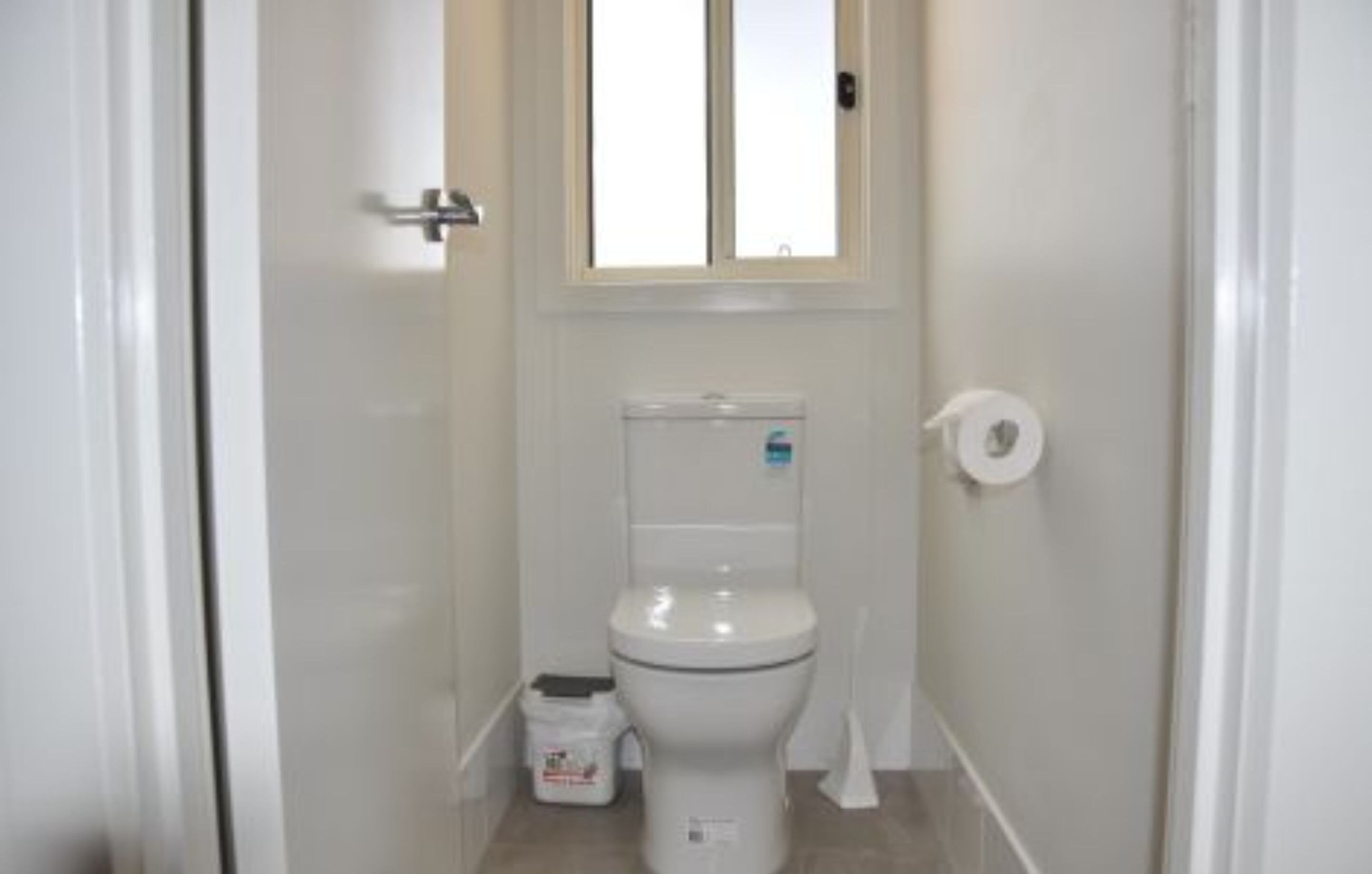 A small, white bathroom with a toilet, a roll of toilet paper, and a trash bin.