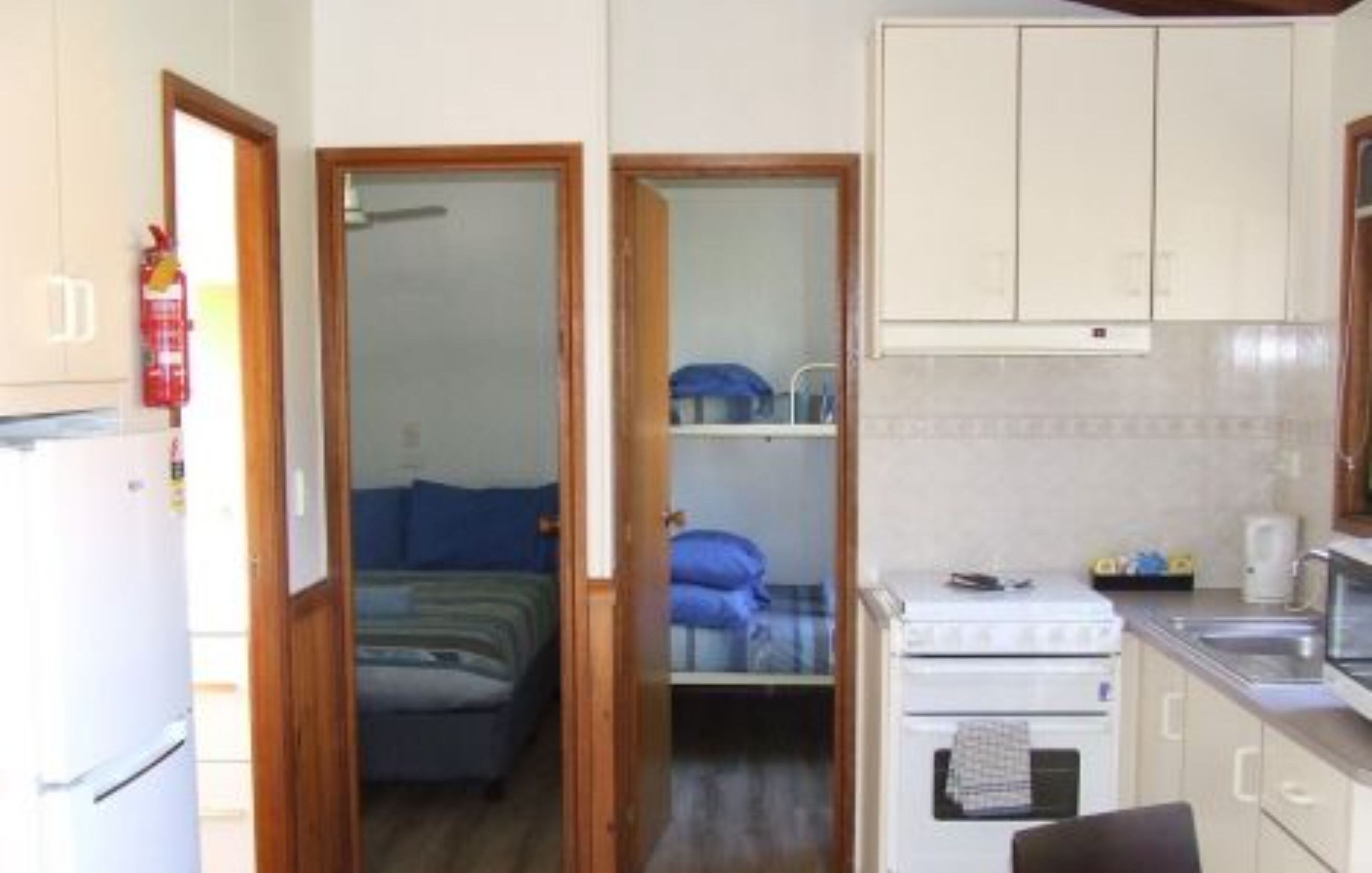 Ein kompakter Raum mit einem Bett, einer kleinen Küchenzeile inklusive Herd und Spüle sowie einem an der Wand befestigten Feuerlöscher.