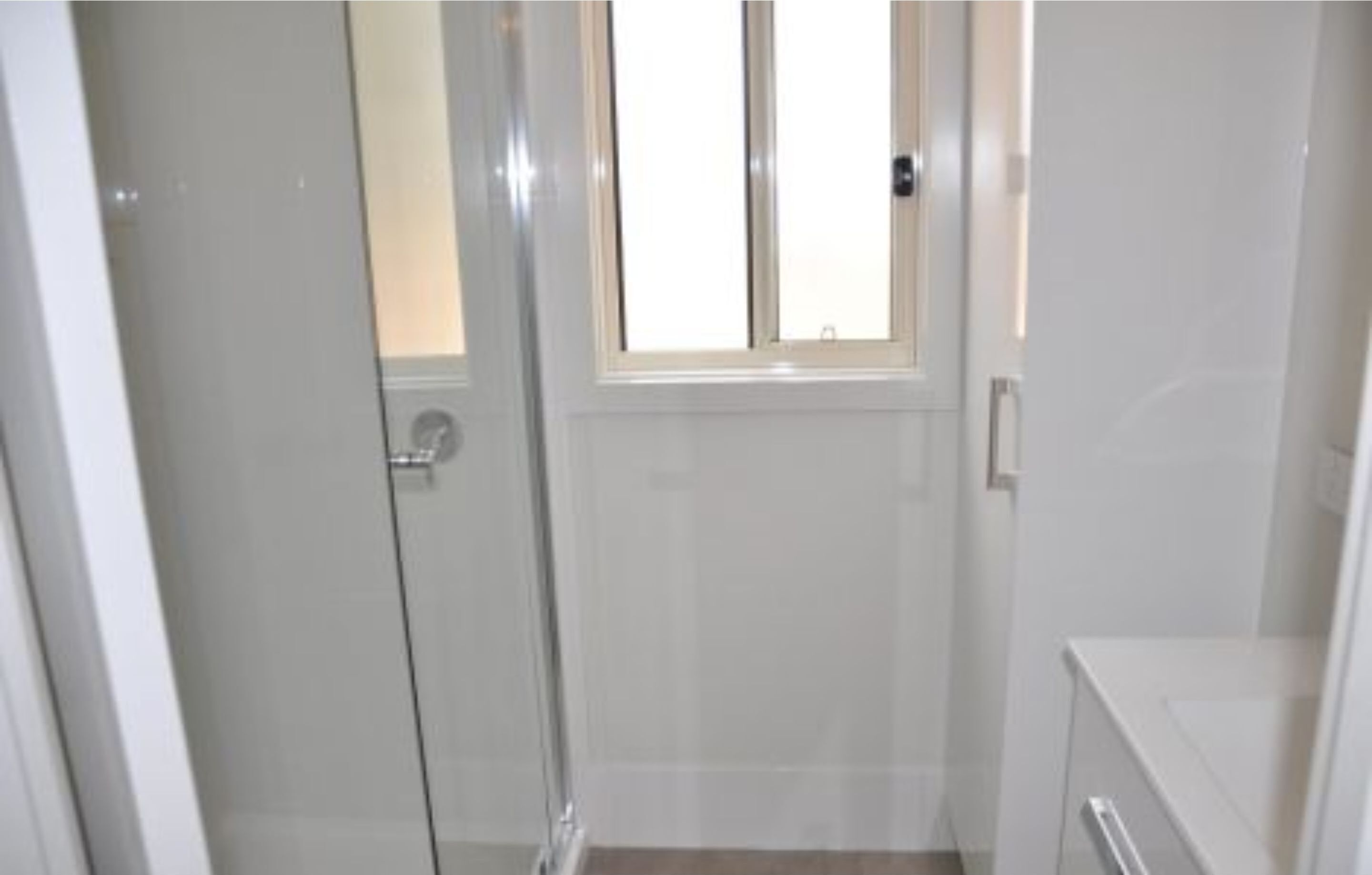 Ein kleines, abgeschlossenes Badezimmer mit Duschabtrennung aus Glas, einem Fenster und einem weißen Waschbecken.