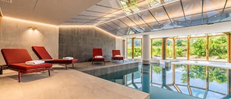 A modern indoor pool area with red lounge chairs, a clear glass ceiling, and a view of greenery outside.