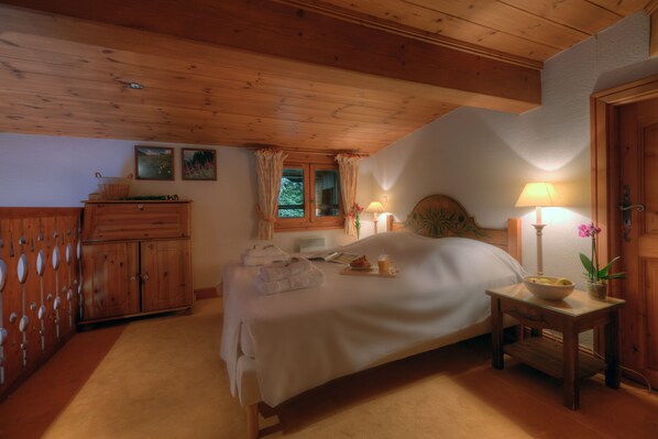 A bedroom with a wooden ceiling, a bed with white linens, a bedside table with a lamp, and a window with curtains.