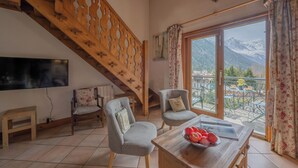 Une pièce avec un escalier en bois, deux fauteuils, une table avec des fruits, un téléviseur et un balcon offrant une vue sur la montagne.