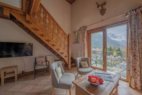 Une pièce avec un escalier en bois, deux fauteuils, une table avec des fruits, un téléviseur et un balcon offrant une vue sur la montagne.