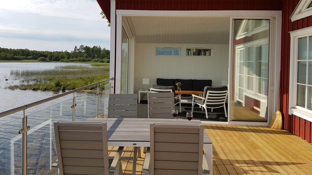 A wooden deck with a table and chairs, a sofa, and a glass railing overlooking a body of water.