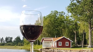A glass of red wine on a lakeside table with a red house in the background.