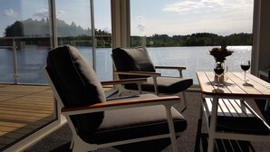 A room with large windows overlooking a lake, featuring two chairs and a table with a vase and glasses.