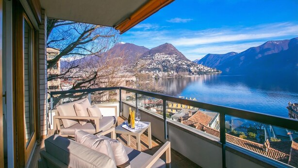 Un balcon avec vue sur un lac et des montagnes, aménagé avec un canapé et des fauteuils, ainsi qu’une table sur laquelle se trouvent une bouteille et des verres.