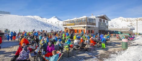 Un espacio al aire libre con mesas y sillas lleno de gente, un edificio con balcón y una montaña nevada al fondo.