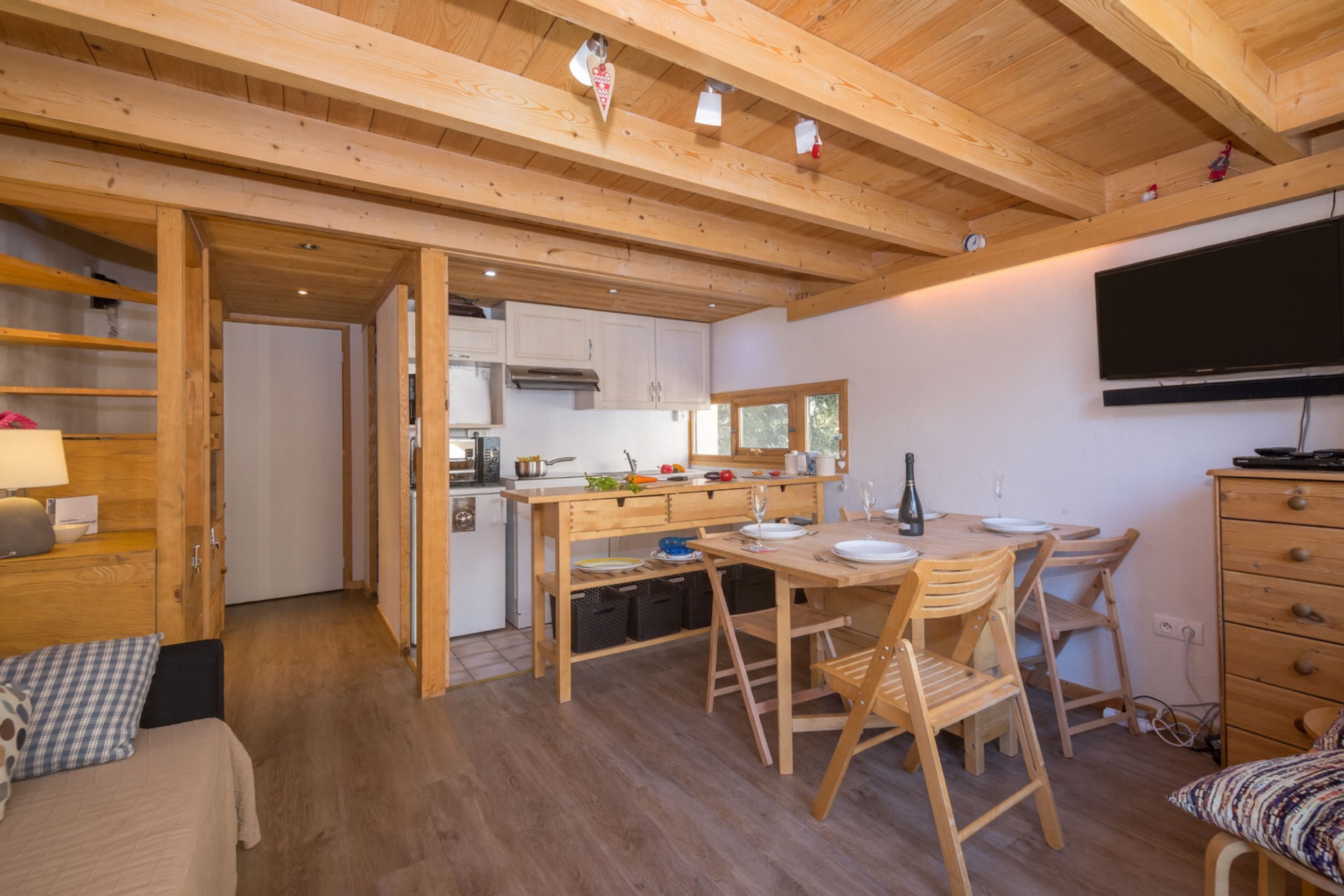 Een knus vertrek met een houten plafond, een eettafel met stoelen, een aan de muur bevestigde televisie en een bed met een gestreept kussen.