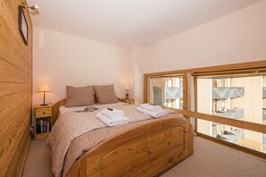 A bedroom with a wooden bed, a nightstand with books, a window with a view of buildings, and a wooden wall. - Grand Roc B220 260m from Slopes, View, Parking & WiFi, Argentière (Chamonix), France (Argentière (Chamonix))