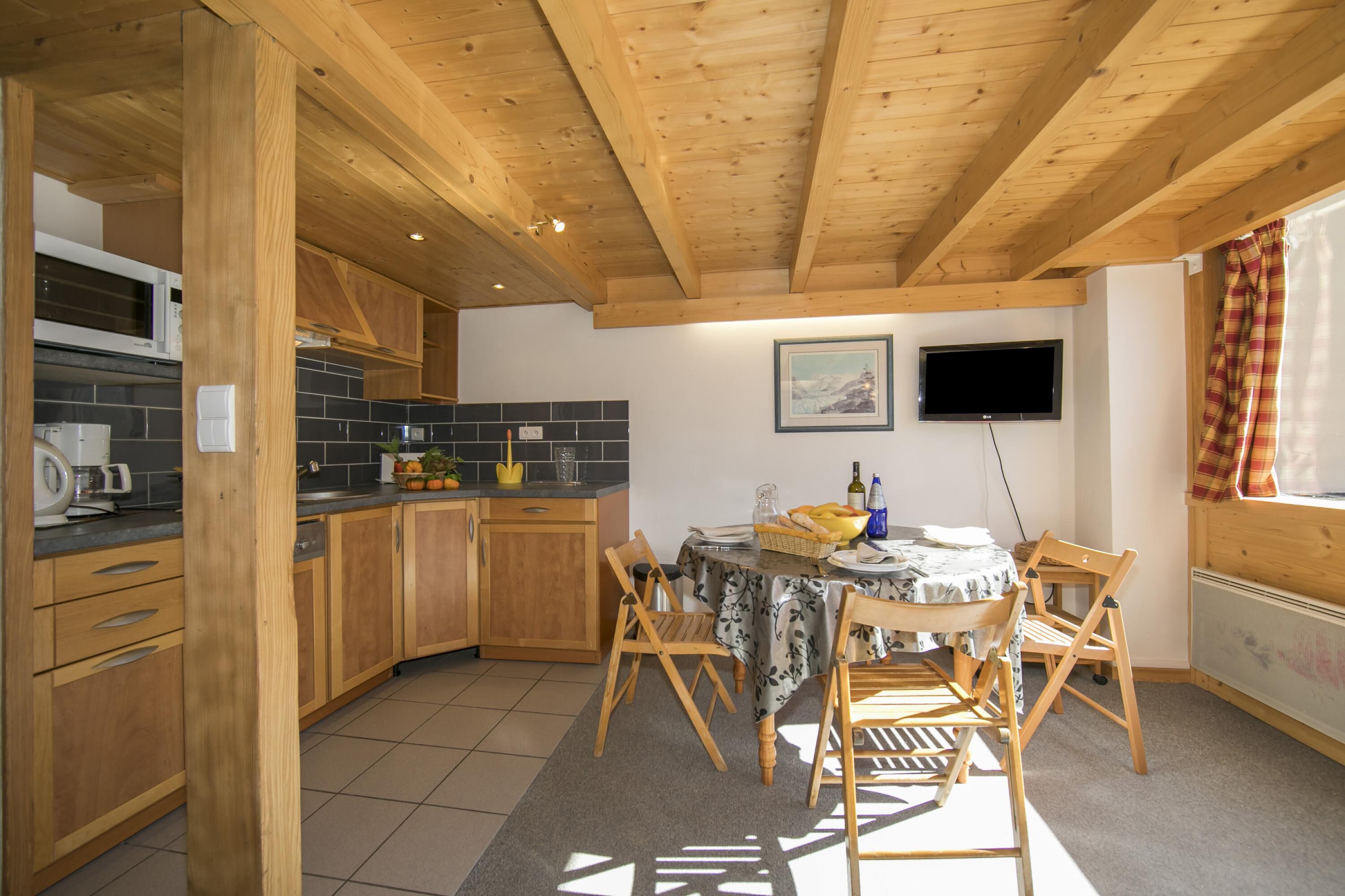Una cocina con armarios de madera, salpicadero de azulejos, una mesa de comedor con sillas y un televisor colgado en la pared.