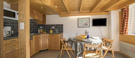 Una cocina con armarios de madera, salpicadero de azulejos, una mesa de comedor con sillas y un televisor colgado en la pared.