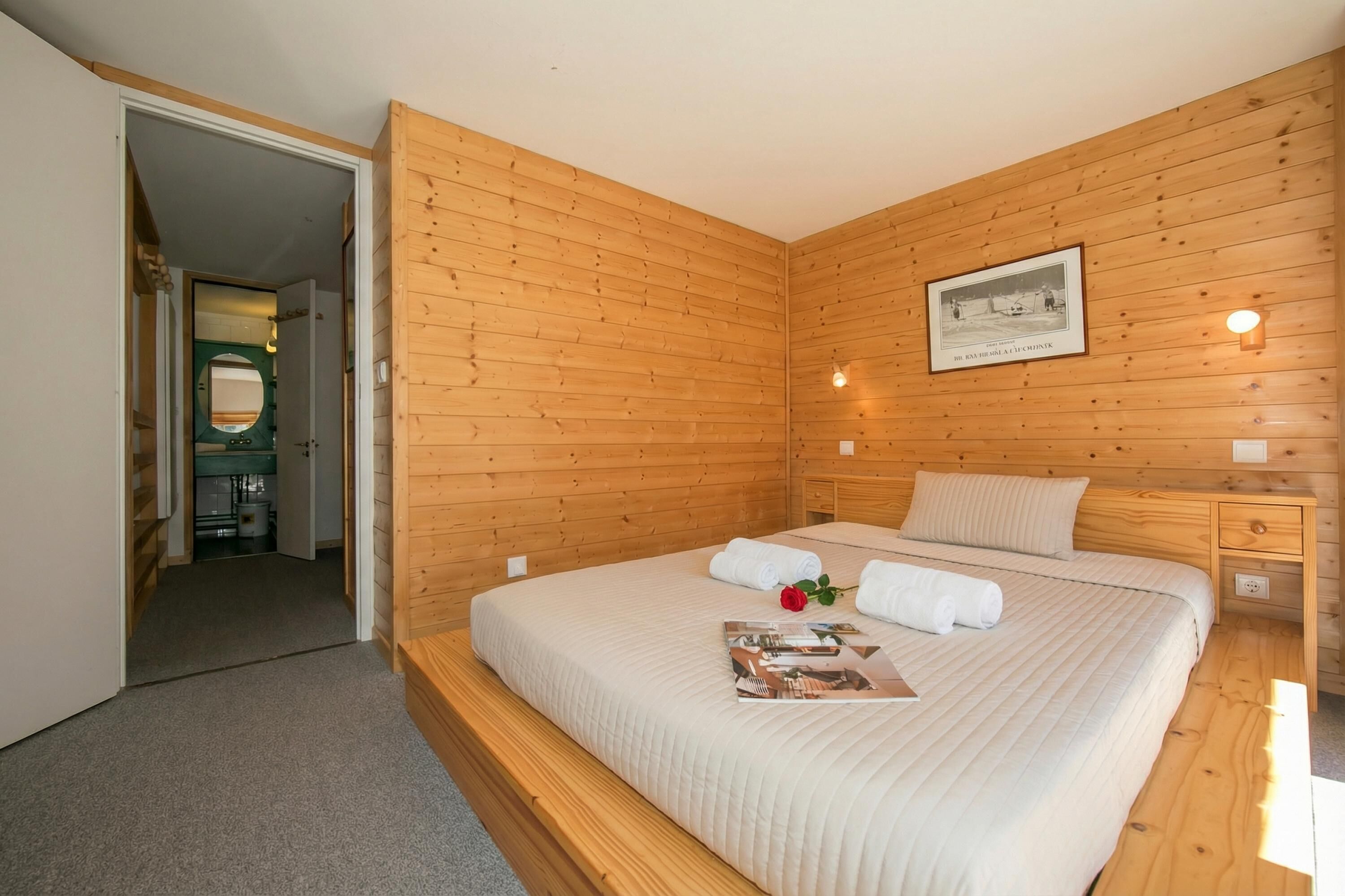 A bedroom with wooden panel walls, a bed with white bedding, a framed picture on the wall, and a small bedside table with a lamp.