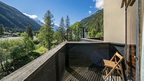 A balcony with a wooden floor and a chair, overlooking a mountainous landscape with trees and a clear sky.