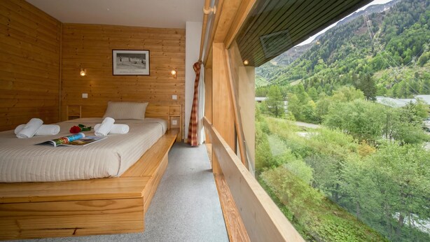 A bedroom with a wooden floor, a bed with a white comforter, a framed picture on the wall, and a view of a green valley through a window.