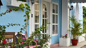 Um edifício azul com janelas brancas e cerca de piquete branca. Há plantas em vasos e uma cadeira rosa na parte externa.