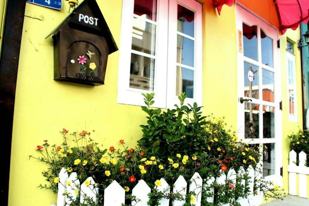 Un edificio amarillo con cerca de piquetes blancos, un comedero de pájaros con la etiqueta 'POST' y un ventanal con toldo rojo.