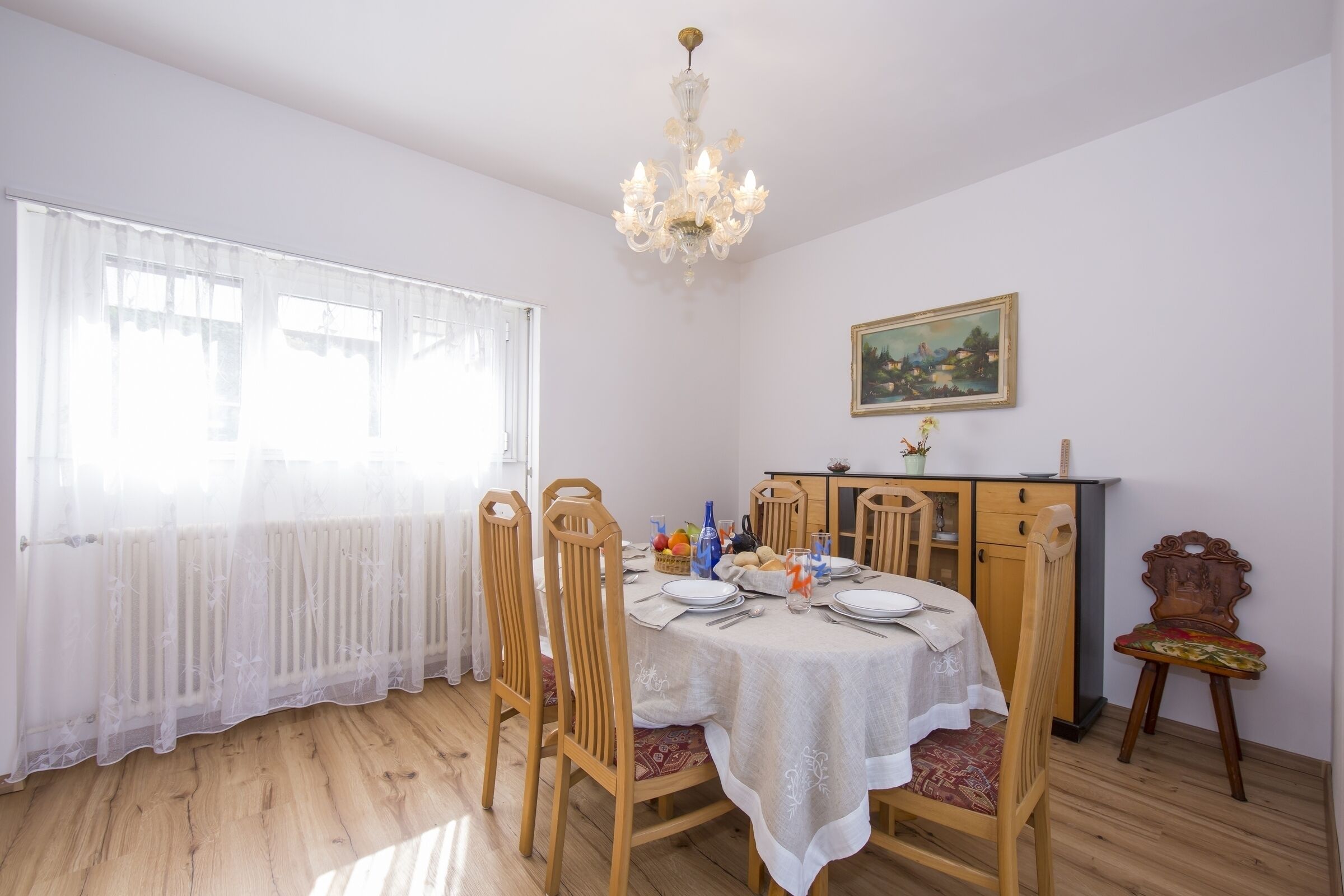 Una sala da pranzo con un tavolo imbandito per quattro persone, sedie in legno, un lampadario, un termosifone e un quadro appeso al muro.