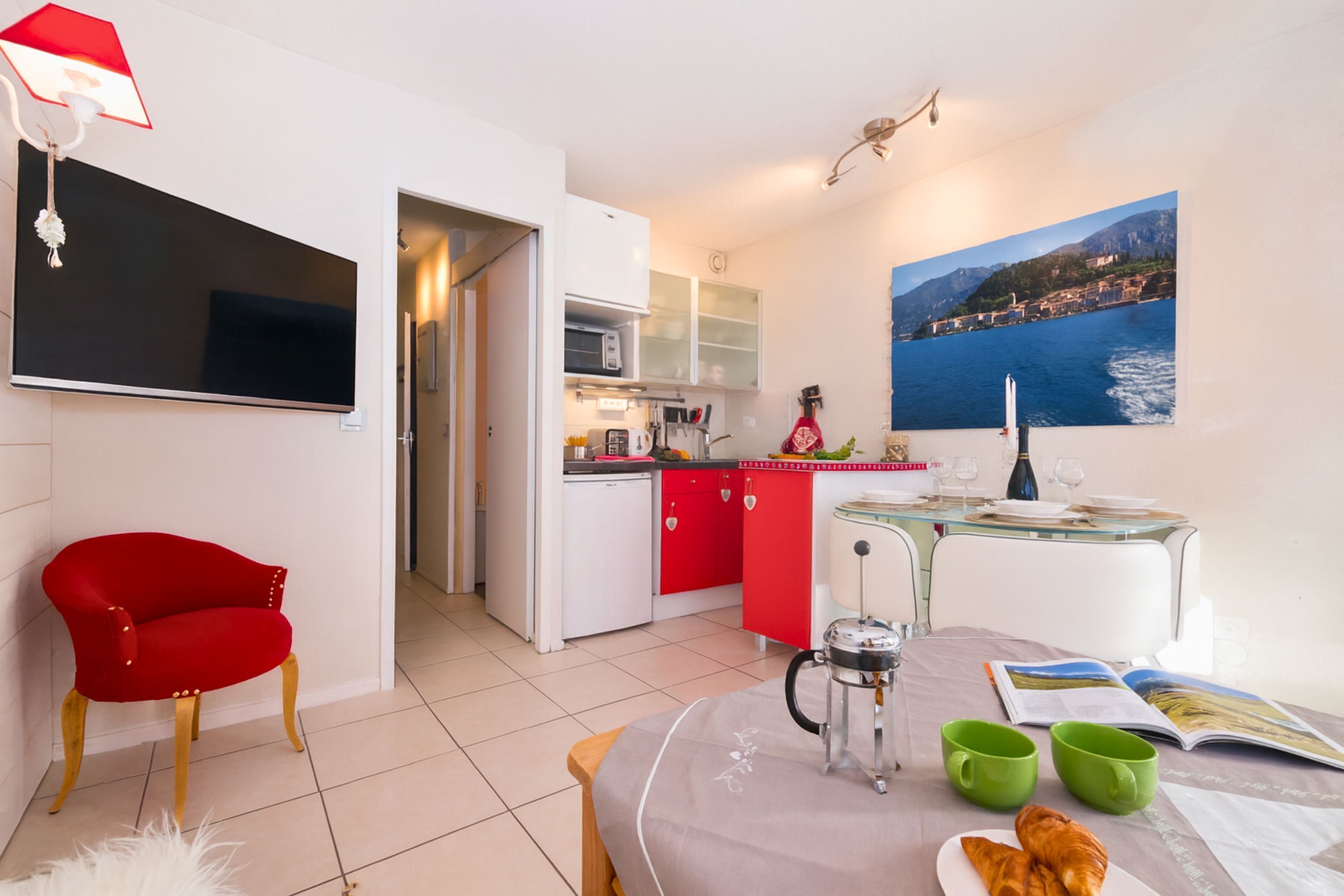 A modern room with a bed, a red chair, a kitchen area, and a dining table with a view of the sea.
