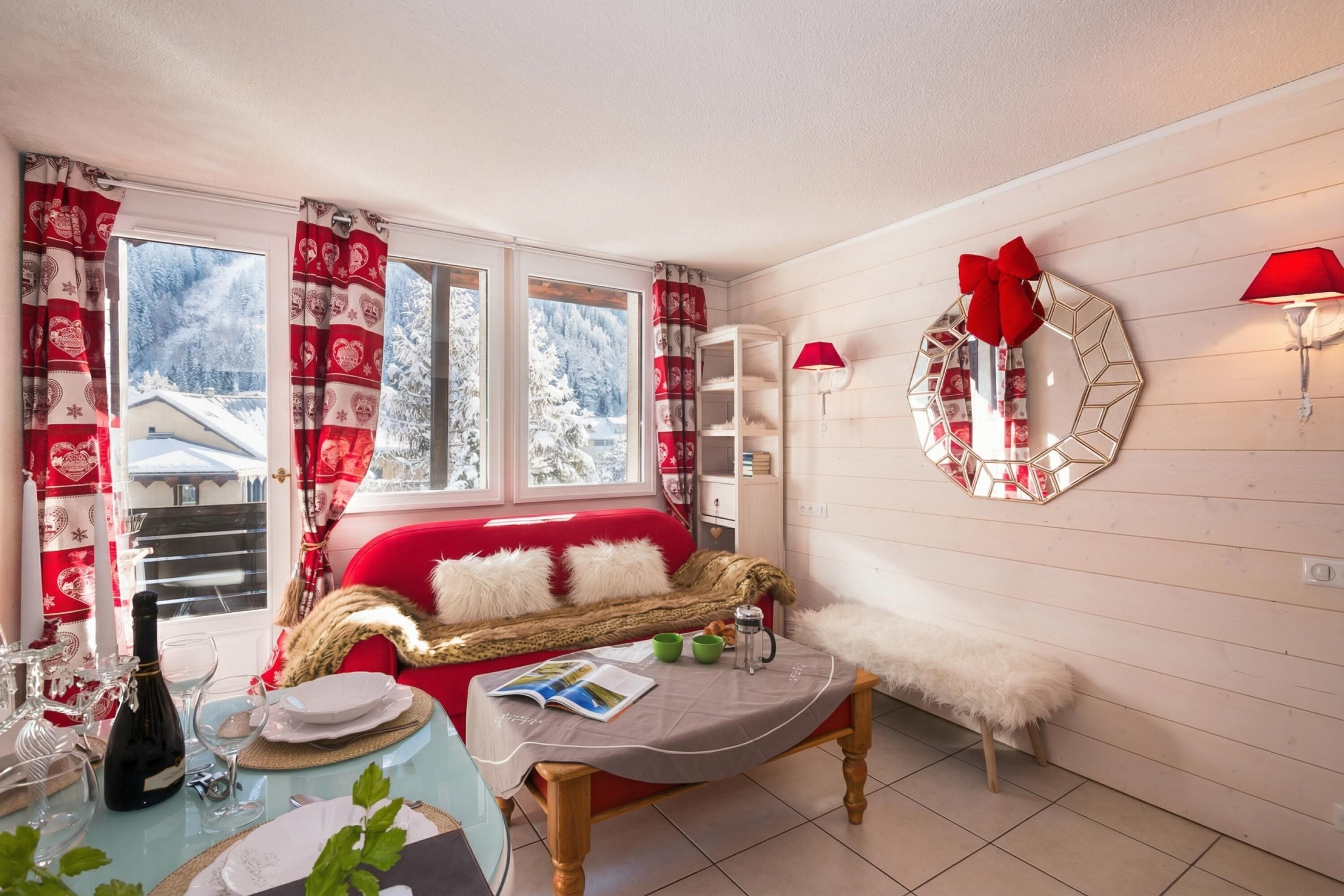 A cozy room with a red sofa, a wooden table with a magazine, a mirror with a bow, and a red lamp.