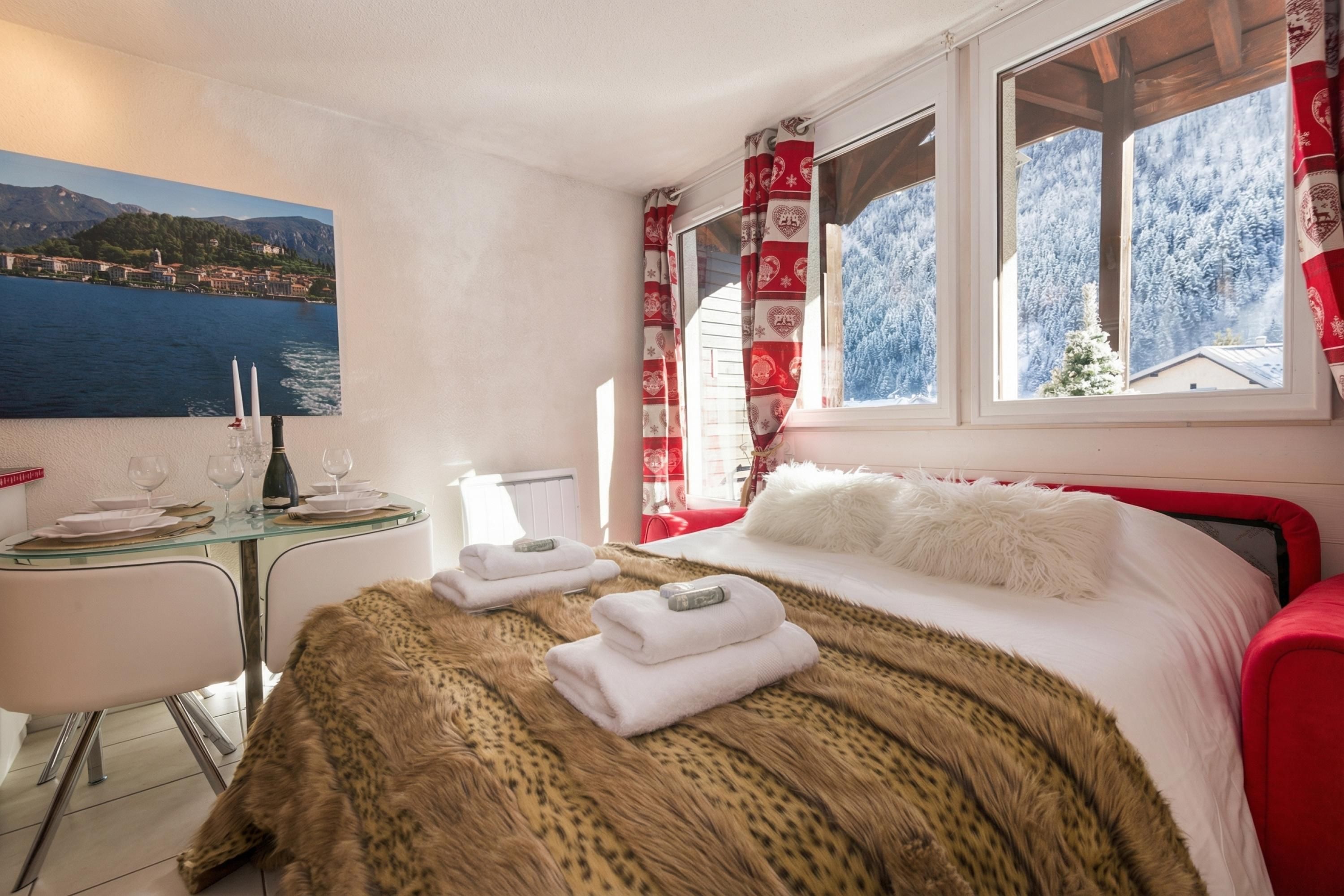 Ein Hotelzimmer mit Bett, einem Esstisch mit Stühlen, einem Bild von einem See und einem Fenster mit Blick auf schneebedeckte Bäume.