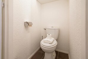 Een kleine badkamer met witte wanden, een wit toilet, een rol toiletpapier en een handdoek op de deksel.